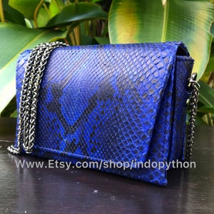 Może przedstawiać: Mała, niebiesko-czarna torebka na ramię ze skóry pytona z srebrnym łańcuszkiem. Torebka ma klapkę i jest wykonana z prawdziwej skóry pytona.