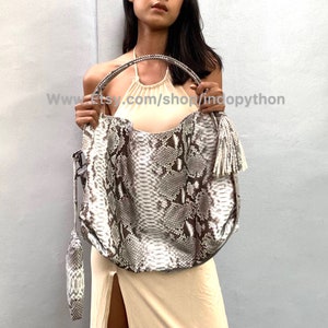 May include: A large, white and brown snakeskin handbag with a tassel. The bag has a top handle and a detachable shoulder strap.