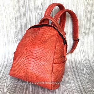 Pu&ograve; includere: Uno zaino arancione brillante in pelle di serpente con chiusura a zip e spallacci regolabili. Lo zaino &egrave; realizzato in vera pelle e presenta un design unico e testurizzato.