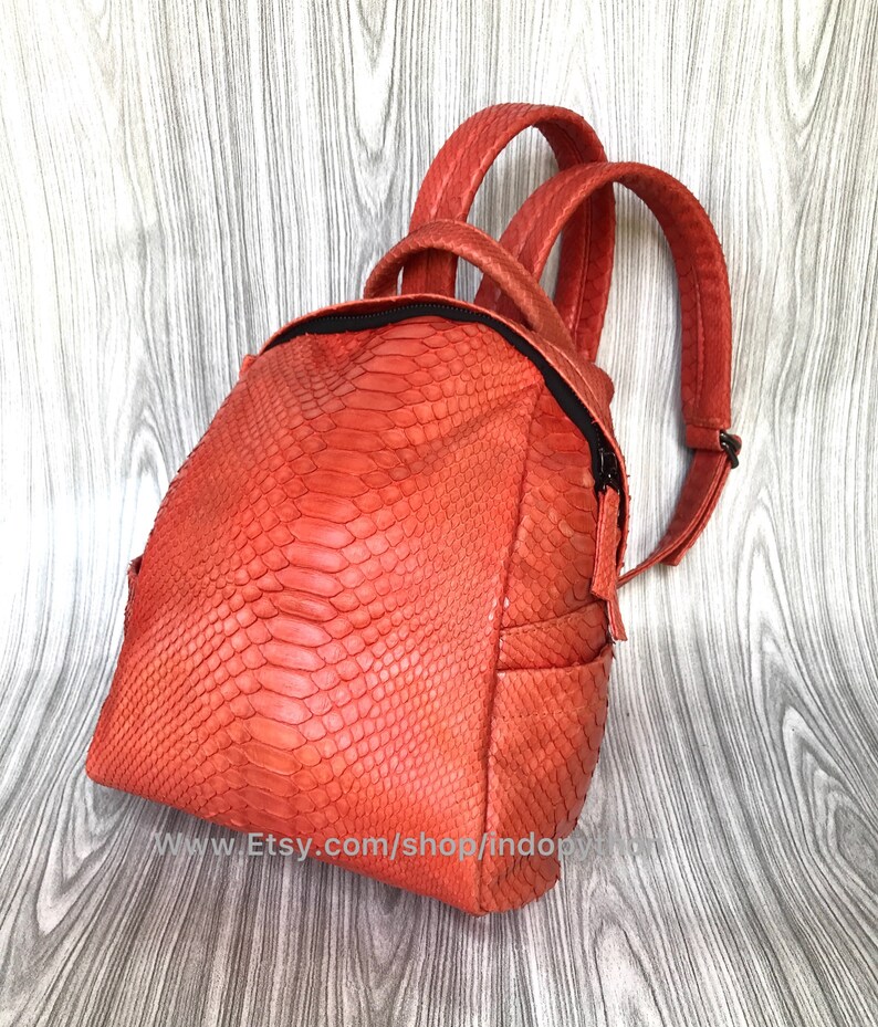 Pu&ograve; includere: Uno zaino arancione brillante in pelle di serpente con chiusura a zip e spallacci regolabili. Lo zaino &egrave; realizzato in vera pelle e presenta un design unico e testurizzato.