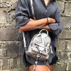 K&ouml;nnte beinhalten: Ein kleiner, grau-wei&szlig;er Schlangenleder-Rucksack mit Rei&szlig;verschluss und Quaste. Der Rucksack wird von einer Person getragen, die ein graues Hemd und schwarze Shorts tr&auml;gt.