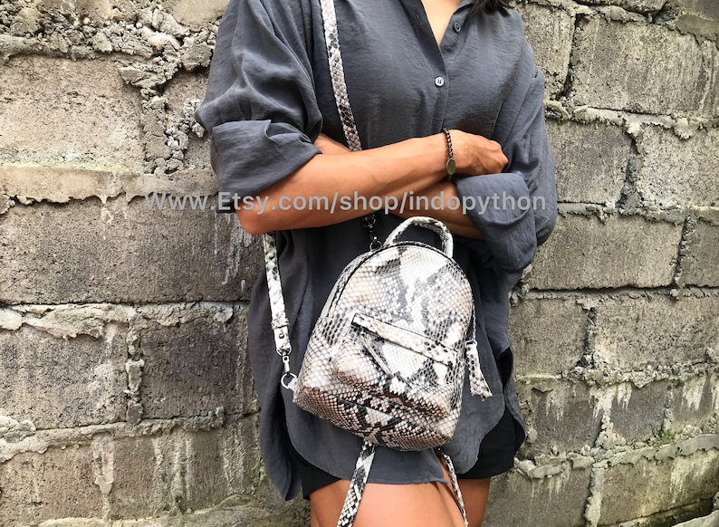 K&ouml;nnte beinhalten: Ein kleiner, grau-wei&szlig;er Schlangenleder-Rucksack mit Rei&szlig;verschluss und Quaste. Der Rucksack wird von einer Person getragen, die ein graues Hemd und schwarze Shorts tr&auml;gt.