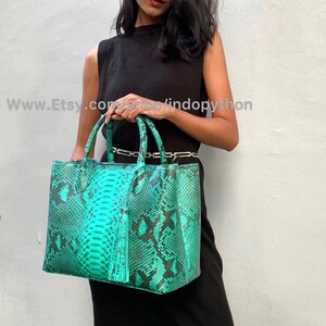 Puede incluir: Un bolso tote de piel de serpiente verde turquesa con un borla negra. El bolso tiene una cremallera superior y dos asas superiores.