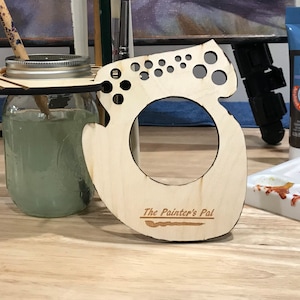 May include: Wooden paintbrush holder with a cutout design and the text "The Painter's Pal". The holder has a circular opening for a jar or cup and multiple small holes for holding paintbrushes.