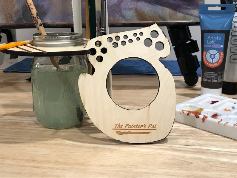 May include: Wooden paintbrush holder with a cutout design and the text "The Painter's Pal". The holder has a circular opening for a jar or cup and multiple small holes for holding paintbrushes.