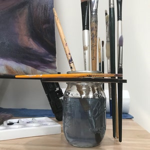 May include: A clear glass jar with a black lid filled with water and paintbrushes. The jar is sitting on a wooden surface with a white paint palette and a canvas in the background.