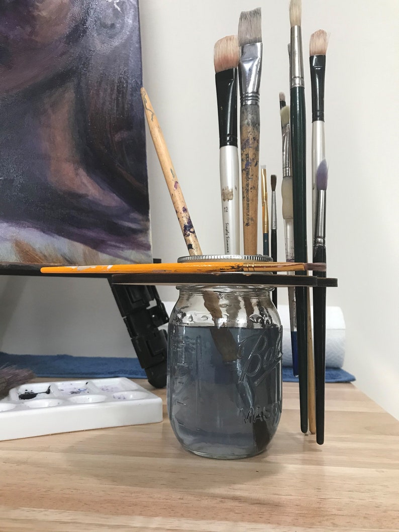 May include: A clear glass jar with a black lid filled with water and paintbrushes. The jar is sitting on a wooden surface with a white paint palette and a canvas in the background.