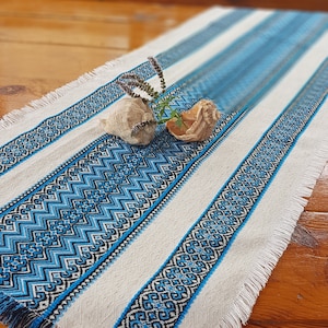 Könnte beinhalten: Ein weißer Tischläufer mit blauen und schwarzen geometrischen Mustern und einem Fransenrand. Der Läufer liegt auf einem Holztisch mit zwei Muscheln und einem Zweig getrockneter Blumen darauf.