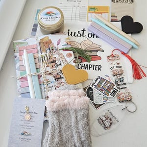 Può includere: Una collezione di articoli a tema libri, tra cui una candela, calzini, adesivi, penne, segnalibri e un portachiavi. Gli articoli sono disposti su una superficie bianca, con la scritta "Cozy Chapter Book Nook" visibile sulla candela.
