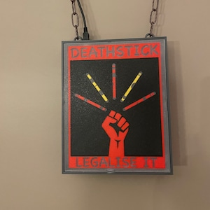 May include: A black and red sign with the text "DEATHSTICK LEGALISE IT" hanging from a chain. The sign features a red fist with five yellow and black sticks radiating from it.