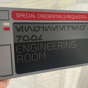 Peut inclure: Une plaque grise et noire avec le texte "SPECIAL CREDENTIALS REQUESTED" en rouge. La plaque comporte également le texte "ENGINEERING ROOM" et une série de symboles et de chiffres.