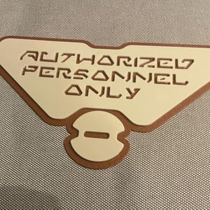 Può includere: Un cartello beige con bordi marroni che recita "AUTHORIZED PERSONNEL ONLY" in un carattere stilizzato.