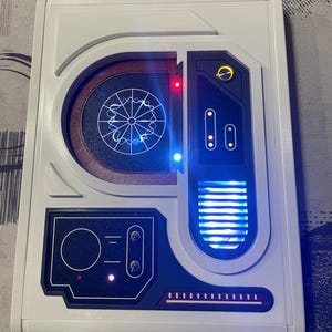 Puede incluir: Un panel de control rectangular blanco con una pantalla circular azul y blanca, un altavoz azul y blanco y un panel negro con tres botones y una luz roja. El panel tiene un borde blanco y un fondo negro.