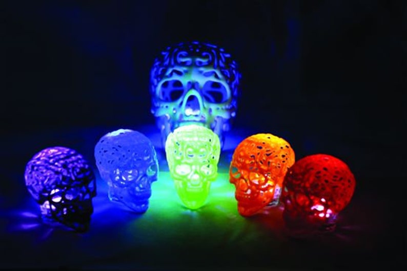 Pu&ograve; includere: Sette piccole figurine di teschio luminose, dal design intricato, in vari colori, tra cui viola, blu, verde, arancione e rosso, disposte su una superficie scura.