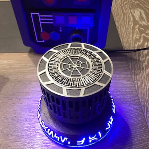 Puede incluir: Un modelo impreso en 3D de una Estrella de la Muerte de Star Wars con un anillo de luz azul alrededor de la base. El anillo tiene una serie de símbolos en blanco.