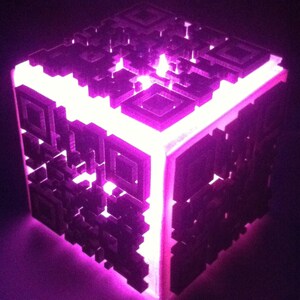 Peut inclure: Une lampe en forme de cube avec une lumière blanche qui brille à travers un motif de code QR noir et blanc sur chaque face. La lampe est faite d'un matériau translucide et est éclairée de l'intérieur, créant un effet lumineux.
