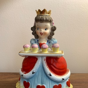 May include: A vintage ceramic figurine of a queen holding a tray of cupcakes. The queen wears a gold crown, a red and blue dress with heart details, and a yellow skirt. The cupcakes have pink frosting.