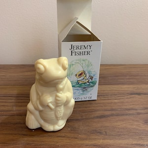 May include: A white ceramic frog figurine holding a fishing rod. The frog is wearing a green vest and a brown hat. The figurine is in a white box with the text "Jeremy Fisher" on it. The box also has a picture of a frog fishing.