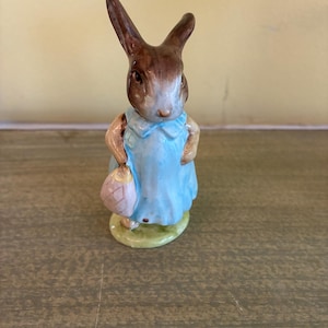 May include: A ceramic figurine of a brown rabbit standing upright. The rabbit wears a light blue collared dress and carries a small, pale pink patterned bag in one hand. The figure is positioned on a small, oval-shaped green base.