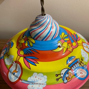 May include: A vintage tin toy top with a red wooden handle and a twisted metal spindle. The top features a colorful design with birds riding bicycles and other whimsical illustrations. The top's body is painted with stripes of red, yellow, and blue.
