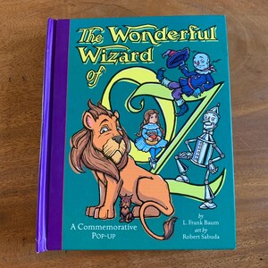 May include: A hardcover book titled "The Wonderful Wizard of Oz" with a teal cover and purple spine. The cover features illustrations of characters from the story, including a lion, Dorothy, the Scarecrow, and the Tin Man. Text includes "A Commemorative POP-UP".