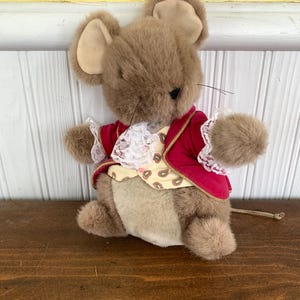 May include: A plush toy mouse with brown fur, large ears, and a long tail. The mouse wears a red velvet jacket with gold trim, a paisley vest, and a white lace collar. The toy is posed on a wooden surface.