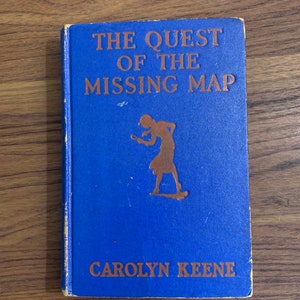 May include: A blue book with the title "The Quest of the Missing Map" and the author "Carolyn Keene" in gold lettering. A silhouette of a girl with a magnifying glass is on the cover.