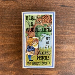May include: A box of six colored pencils featuring an illustration from "Alice in Wonderland." The box has a blue border and includes images of Alice, the Cheshire Cat, and text that reads "For Artists Over 3."