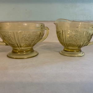 May include: A vintage yellow glass sugar bowl and creamer set with a floral design. The set has a footed base and two handles on the sugar bowl.