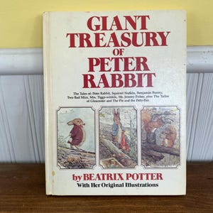 May include: A vintage copy of "Giant Treasury of Peter Rabbit" by Beatrix Potter. The book cover features the title in red lettering, with illustrations of Peter Rabbit and other characters. The book is on a wooden surface.