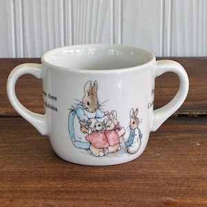 May include: White ceramic mug with two handles, featuring illustrations of rabbits in various outfits. The mug has text on the side. The design includes a rabbit wearing a blue jacket and a pink dress. The mug is sitting on a wooden surface.
