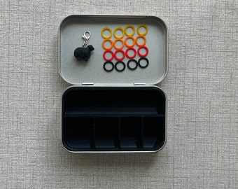 May include: A silver metal box with a black sheep charm and a compartment for storing small items. The box has a clear lid with a grid of colorful rubber rings.