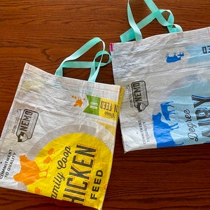 May include: Two reusable shopping bags made from recycled plastic. The bag on the left is yellow and white with the text "Family Coop Chicken Feed" and a picture of a chicken. The bag on the right is blue and white with the text "Degree Airy" and a picture of a cow. Both bags have turquoise handles.