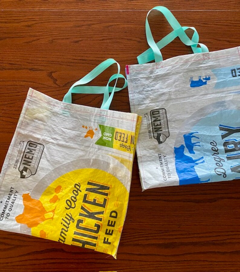 May include: Two reusable shopping bags made from recycled plastic. The bag on the left is yellow and white with the text "Family Coop Chicken Feed" and a picture of a chicken. The bag on the right is blue and white with the text "Degree Airy" and a picture of a cow. Both bags have turquoise handles.