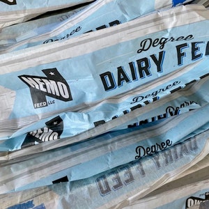 May include: Close-up of several blue and white bags of Degree Dairy Feed. The bags are stacked on top of each other and the text "Degree Dairy Feed" is visible on the front of each bag.