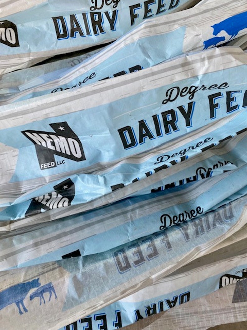 May include: Close-up of several blue and white bags of Degree Dairy Feed. The bags are stacked on top of each other and the text "Degree Dairy Feed" is visible on the front of each bag.