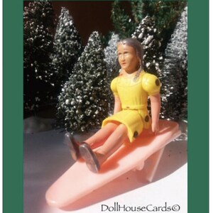 May include: A yellow dress with black polka dots is worn by a doll sitting on a pink plastic sled. The doll has brown hair and a light complexion. The doll is sitting in a snowy forest setting.
