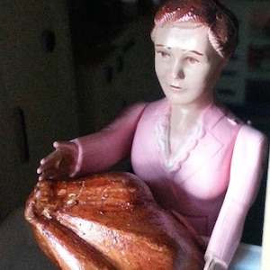 May include: A dollhouse figurine of a woman in a pink dress holding a brown roasted turkey.