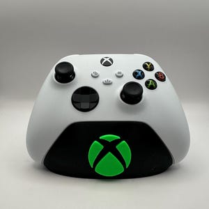 May include: A white Xbox controller is sitting on a black charging stand with a green Xbox logo. The controller has black buttons and a black D-pad.