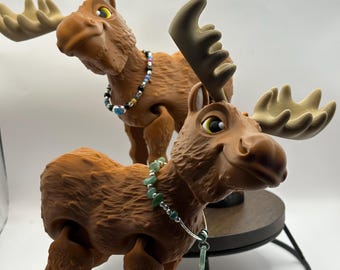 May include: Two brown moose figurines with articulated legs and antlers. The moose on the left is wearing a multi-colored beaded necklace. The moose on the right is wearing a necklace of green gemstone beads.