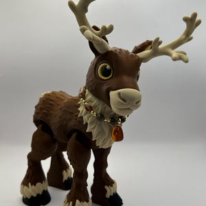 May include: A brown reindeer figurine with large, expressive eyes and tan antlers. The reindeer has a fluffy, textured body and wears a beaded necklace with a pendant. The hooves are black and white. The figurine is standing on a white surface.