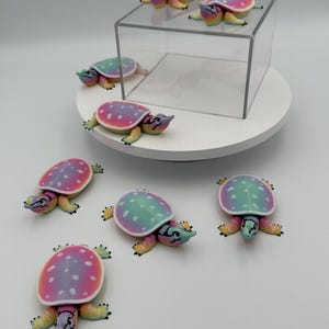 May include: Seven small turtle figurines featuring a gradient shell pattern in shades of pink, purple, and teal with white polka dots. The turtles have yellow limbs and heads with black eye markings. Two turtles are positioned on a clear acrylic cube.