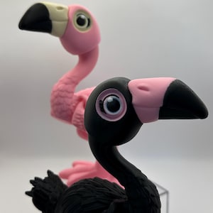 May include: Two flamingo figurines, one pink and one black, are displayed against a white background. The pink flamingo is in the background, while the black flamingo is in the foreground. Both have pink beaks and large, round eyes.