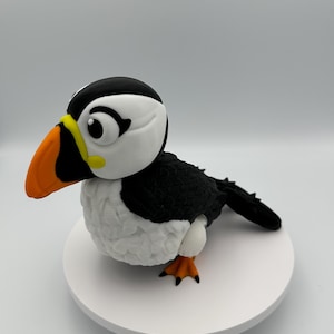May include: A handcrafted puffin figurine with a black back, white belly, and a large orange and yellow beak. The bird has black and white markings on its head and is perched on a white circular base. The puffin's feet are orange.