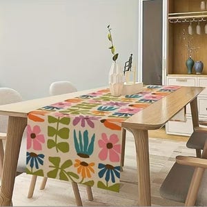 May include: A colorful floral table runner with a repeating pattern of stylized flowers and leaves in pink, orange, blue, and green on a cream background. The runner is displayed on a light wood table, adding a decorative touch to the dining area.