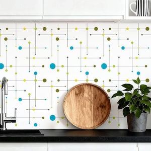 May include: A kitchen backsplash featuring a white backdrop with a repeating pattern of lines and circles in teal, olive green, and grey. A wooden plate, a potted plant, and a coffee machine are on the countertop.