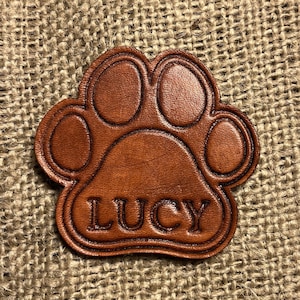 May include: A brown leather paw print with the name "LUCY" embossed in the center.
