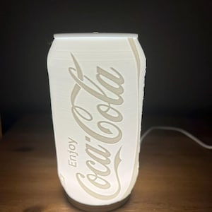 May include: A white, illuminated lamp shaped like a Coca-Cola can. The lamp features the Coca-Cola logo and the word "Enjoy" in a stylized font. The lamp is plugged in and sits on a wooden surface.