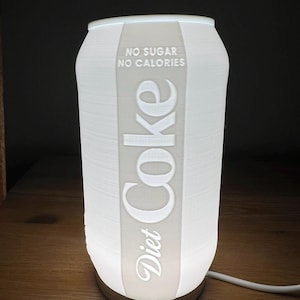 May include: A white Diet Coke can-shaped lamp with the words "Diet Coke", "NO SUGAR", and "NO CALORIES" printed on the front. The lamp is illuminated, and the base is a light beige color. A white cord extends from the base.