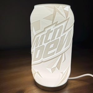 May include: A white, cylindrical lamp shaped like a Mountain Dew soda can. The lamp features the "Mtn Dew" logo and geometric patterns. The lamp is lit from within, and sits on a wooden surface.